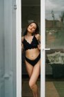 Content slim female in black lingerie standing near glass door leading to balcony and looking away — Stock Photo