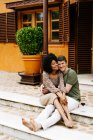 Позитивная многорасовая пара, сидящая на каменной лестнице крыльца коттеджа и обнимающаяся, наслаждаясь выходными вместе — стоковое фото