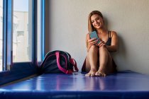 Atleta femenina alegre sentada en la estera con bolsa de deporte en el gimnasio y el teléfono móvil de navegación - foto de stock