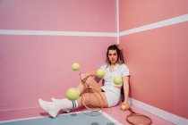 Молодая спортсменка в кроссовках и спортивной одежде сидит с теннисными ракетками и смотрит в сторону от движущихся мячей — стоковое фото