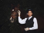 Senhora adulta afro-americana confiante em roupas elegantes e chapéu de pé com cavalo marrom enquanto olha para a câmera perto de árvores durante o dia — Fotografia de Stock