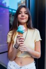 Crop cheerful young female in pendant and earrings with delicious gelato in waffle cone looking at camera on street — Stock Photo