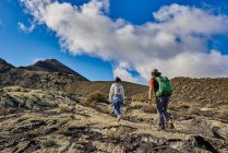 From below man and woman with backpacks walking on rough slope of mountain against cloudy blue sky in Fuerteventura, Spain — Stock Photo