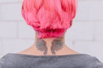 Vue de dos récolte femelle méconnaissable avec des cheveux teints et tatouage des ailes sur le cou debout près du mur blanc — Photo de stock