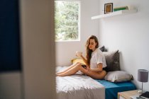 Side view of happy female in panties and t shirt surfing Internet on cellphone while sitting on comfortable bed with cup of drink and enjoying morning — Stock Photo
