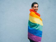 Gay etnico maschio avvolto in arcobaleno LGBT bandiera guardando fotocamera contro grigio muro in città — Foto stock