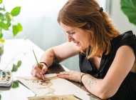 Umtriebige Designerin malt mit Pinsel auf Papier, während sie im Kreativbüro am Tisch sitzt und an einem Projekt arbeitet — Stockfoto