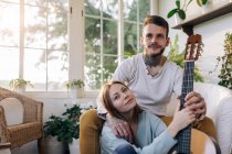 Content female Cheerful tattooed male musician playing guitar near Cheerful tattooed male musician while looking at each other in armchair in house room — Stock Photo