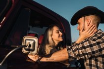 Side view of delighted woman sticking out of vintage car window and embracing boyfriend in plaid shirt and cowboy hat at sunset in countryside — Stock Photo