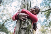 De baixo de criança étnica encantadora tocando casca áspera de tronco de árvore envelhecida com líquen enquanto olha para a câmera na floresta — Fotografia de Stock