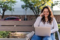 Jovem e alegre estudante latino-americana conversando no celular e navegando no laptop enquanto descansa no banco de madeira na rua da cidade no dia de verão — Fotografia de Stock