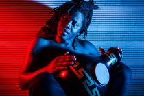 Verträumter schwarzer Musiker mit nacktem Oberkörper spielt afrikanische Trommel im Studio mit roten und blauen Neonlichtern — Stockfoto