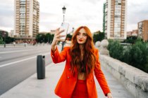 Delighted trendy female with red hair and in orange suit walking along street in city and taking self portrait on smartphone — Stock Photo