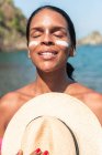 Turista femenina étnica soñadora con protector solar en las mejillas y la nariz de pie con los ojos cerrados y sombrero contra el mar a la luz del sol - foto de stock