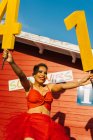 Trendy femmina nera con numeri decorativi e palloncini rossi in piedi sulla passerella contro la costruzione durante la festa di compleanno alla luce del sole — Foto stock