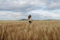 Анонимная женщина с летящими волосами, бегущая по лугу с пшеничными шипами под облачным небом — стоковое фото