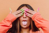 Mulher impertinente mostrando língua enquanto cobre os olhos com as mãos com longas unhas brilhantes contra o fundo laranja — Fotografia de Stock