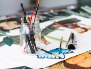 Vaso di vetro con pennelli assortiti posizionato su vari dipinti creativi sul tavolo in officina — Foto stock