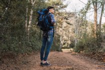 Side view of traveling man with backpack standing on sandy road in forest during trekking and looking away — Stock Photo