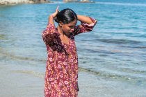 Mulher viajante étnica em vestido de verão com ornamento floral olhando para baixo com as mãos atrás da cabeça contra o oceano e a rocha — Fotografia de Stock