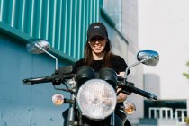 Positive female motorcyclist sitting on motorbike parked in city street on sunny day and looking at camera — Stock Photo