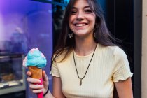 Cosecha alegre joven hembra en colgante y pendientes con delicioso helado en cono de gofre mirando a la cámara en la calle - foto de stock