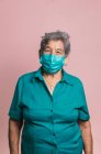 Улыбающаяся пожилая женщина с помощью синей защитной медицинской маски от коронавируса, глядя на камеру на розовом фоне в студии — стоковое фото