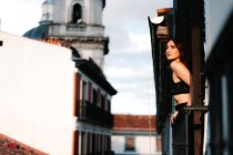 Вид збоку безтурботний гарненька жінка насолоджується сонячним світлом, дивлячись далеко, спираючись на балкони житлового будинку в місті — стокове фото