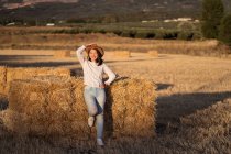 Conteúdo fêmea étnica com chapéu de palha em pé perto da pilha de feno seco no campo e olhando para a câmera — Fotografia de Stock