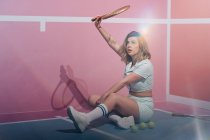 Young sportswoman in sneakers and sports clothes sitting with tennis rackets while looking away — Stock Photo