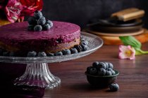 Delicioso pastel de mousse de arándanos con crema púrpura decorada con bayas frescas servidas en soporte de vidrio sobre mesa oscura con flores - foto de stock