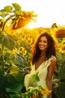Graciosa jovem hispânica fêmea em elegante vestido amarelo de pé em meio a girassóis florescendo no campo rural no dia ensolarado de verão olhando para a câmera — Fotografia de Stock