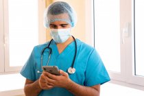 Médico masculino de uniforme e com estetoscópio em pé no hospital e navegar na Internet no smartphone — Fotografia de Stock
