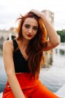 Pretty female with long ginger hair and in bright orange pants sitting on border on promenade in city looking at camera — Stock Photo