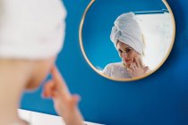 Зворотний вигляд жінки в рушнику, що відбивається у дзеркалі у ванній кімнаті та нанесення крему для обличчя під час процедури догляду за шкірою вранці — стокове фото