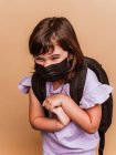 Criança encantada com mochila e máscara protetora de coronavírus em fundo marrom no estúdio — Fotografia de Stock