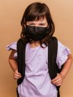 Entzücktes Kind mit Rucksack und Schutzmaske vor Coronavirus auf braunem Hintergrund im Atelier — Stockfoto