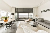 Interior de la moderna y espaciosa sala de estar con cómodo sofá y mesa de madera y sillas en un lujoso apartamento contemporáneo con ventanas panorámicas - foto de stock