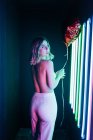 Back view of young sensual woman in pants with balloon looking at camera over shoulder against neon lamps — Stock Photo