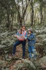 Menina étnica com guia de papel olhando para longe contra o irmão com binóculos entre plantas de samambaia em florestas de verão — Fotografia de Stock