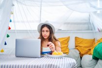 Joven hembra sentada en la mesa y navegando por Internet en netbook mientras come cereza fresca y disfruta del fin de semana de verano en la tienda del patio trasero - foto de stock