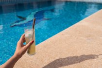 Сверху урожая женщина со стаканом освежающего коктейля с кубиками льда и нулевой металлической соломой, помещенной у бассейна в солнечный день летом — стоковое фото