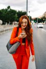 Donna sorridente con i capelli rossi e in tuta arancione che registra messaggi audio sul cellulare mentre comunica con un amico sui social media e cammina per strada — Foto stock