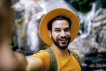 Deleitado viajero masculino en traje amarillo tomando selfie sobre fondo arrugado de rocas durante el trekking en bosques - foto de stock
