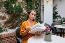 Sereno concurso feminino leitura livro interessante enquanto sentado à mesa no terraço de verão da casa — Fotografia de Stock