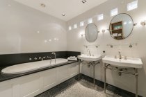 Стильний дизайн інтер'єру будинку просторої світло-білої ванної кімнати з дзеркалами над двома умивальниками та ванною в сучасній квартирі — стокове фото