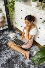 Femme ethnique contemplative avec netbook et verre d'eau reposant sur un tapis doux tout en regardant loin contre miroir dans la maison — Photo de stock