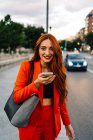 Smiling female with red hair and in orange suit recording audio message on mobile phone while communicating with friend on social media and standing in city street — Stock Photo