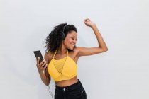 Happy African American femelle dans les écouteurs avec téléphone portable dansant tout en écoutant la chanson sur fond de lumière — Photo de stock