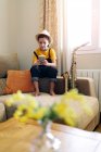 Child in hat text messaging on cellphone while sitting on couch with saxophone in living room — Stock Photo